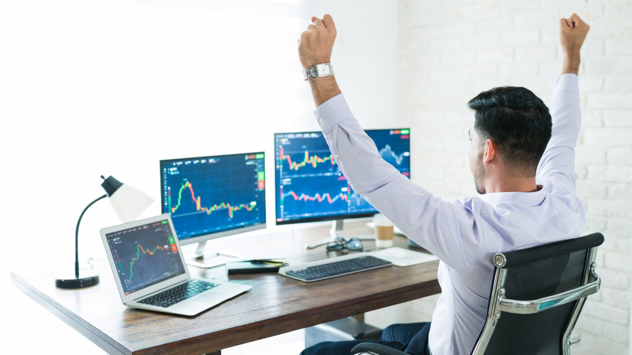 Trader raising both hands, focused on three screens displaying trading charts, illustrating the use of the Slow Stochastic Oscillator for day trading.