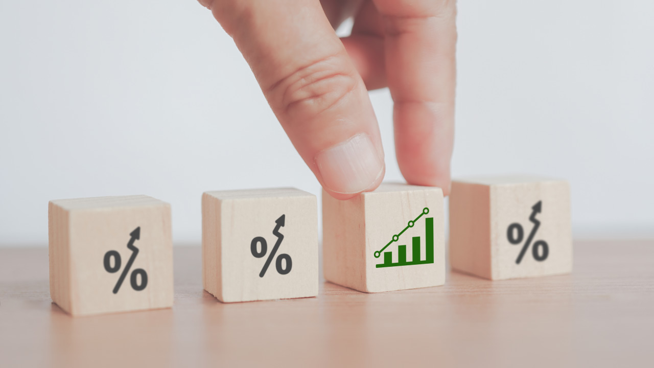 A hand picking up a wooden block displaying a green upward-trending bar chart among blocks with percentage signs and upward arrows, illustrating "How to Trade Low Volatility Markets?"
