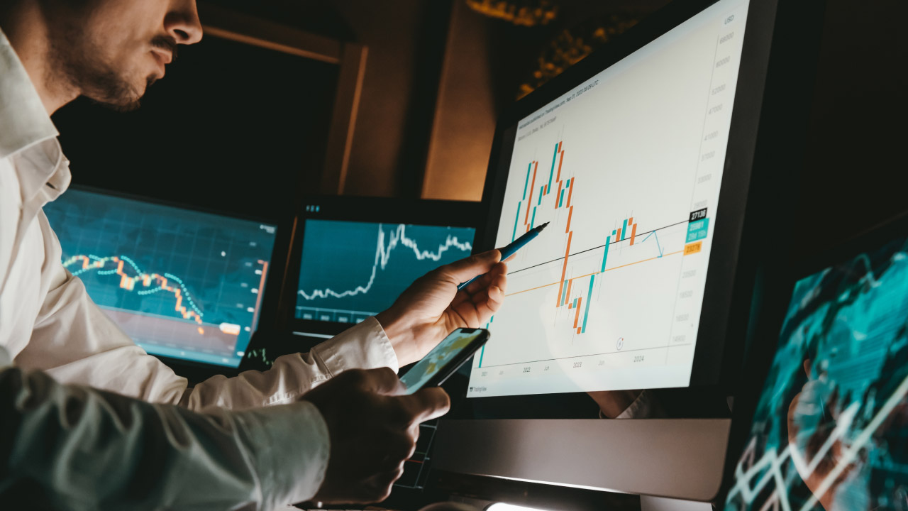  a trader analyzing candlestick chart patterns on a computer monitor while holding a smartphone and a pen