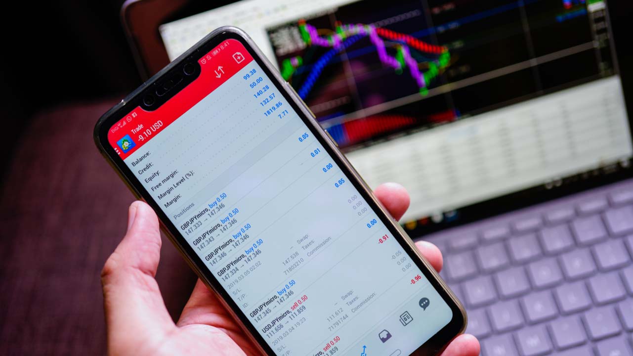 How To Set Up MetaTrader? Step-By-Step Instruction — a person holding a smartphone displaying a trading app with a live account balance, illustrating the MetaTrader setup process.