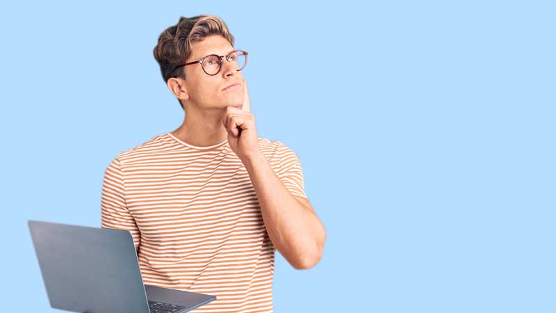 Man wearing glasses and a striped shirt holding a laptop and thinking, standing against a light blue background.