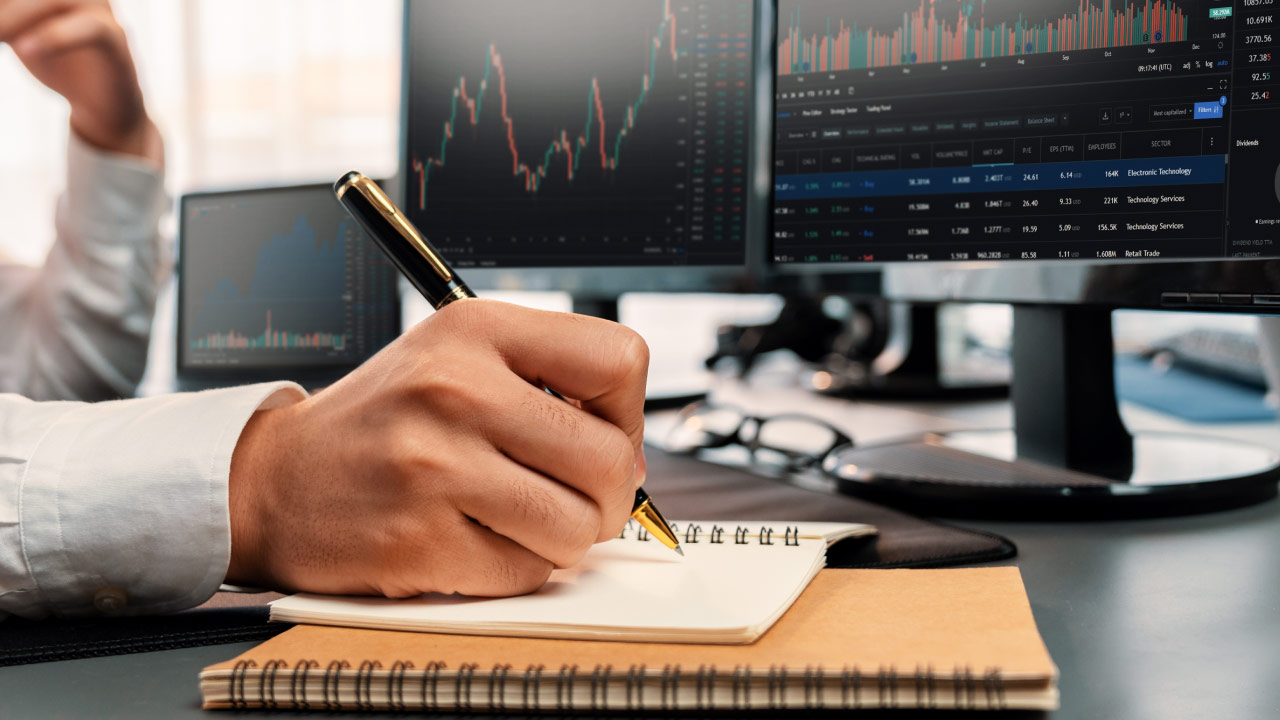 How to Create a Trading Strategy, a person’s hand writing in a spiral notebook with a gold pen, seated at a desk with multiple monitors displaying financial trading charts and stock market data.