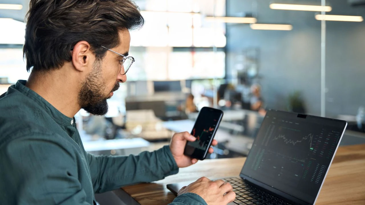Trader holding a phone in front of a laptop, analyzing forex charts to understand ascending and descending triangle patterns.
