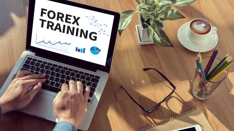 Person using a laptop displaying 'Forex Training' on the screen, with a coffee cup, plant, and stationery on a wooden desk.