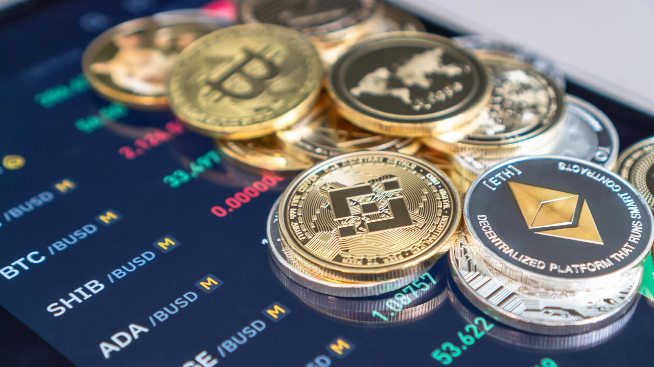 A stack of cryptocurrency coins, including Bitcoin, Ethereum, and Binance Coin, placed on a trading screen displaying market data. Examining the pros and cons of weekend crypto trading.