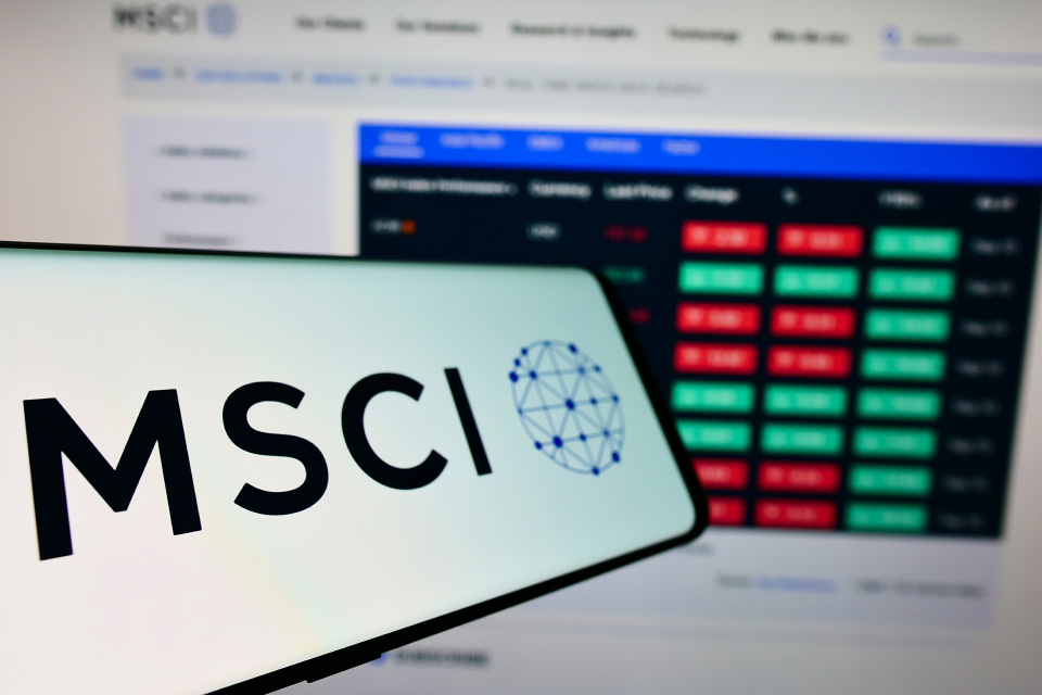 Smartphone displaying the MSCI logo with a blurred computer screen in the background, symbolizing trading the MSCI World Index.