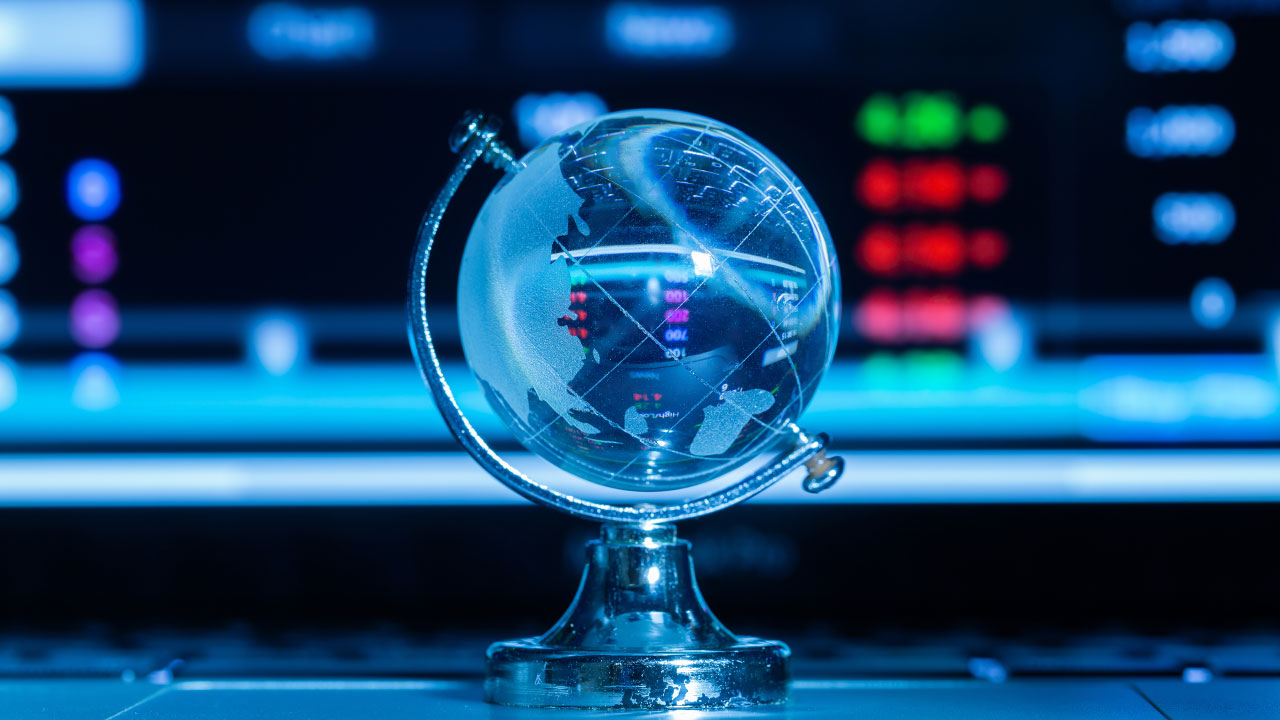 A crystal globe on a stand placed in front of a blurred digital screen displaying market data, illustrating "How to Trade the Impact of Politics on Forex Markets?