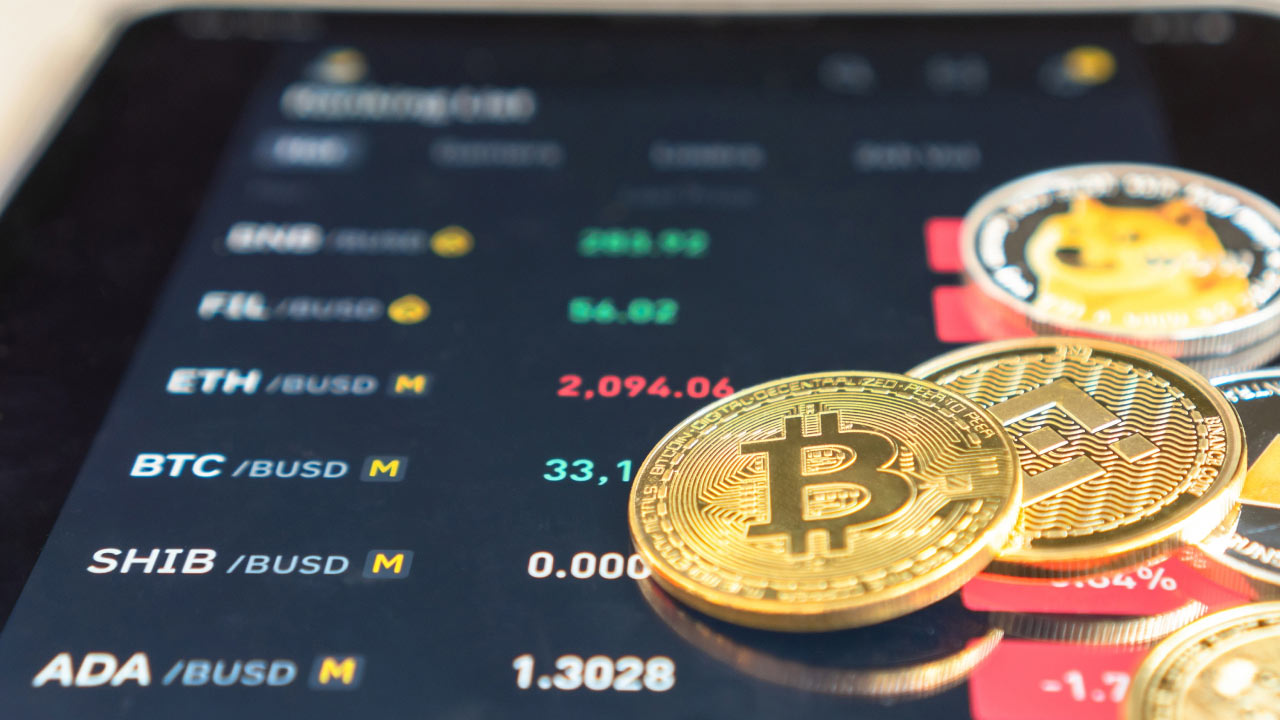 Bitcoin coin in front of a trading monitor displaying cryptocurrency charts, illustrating "How To Cautiously Trade Cryptocurrencies?