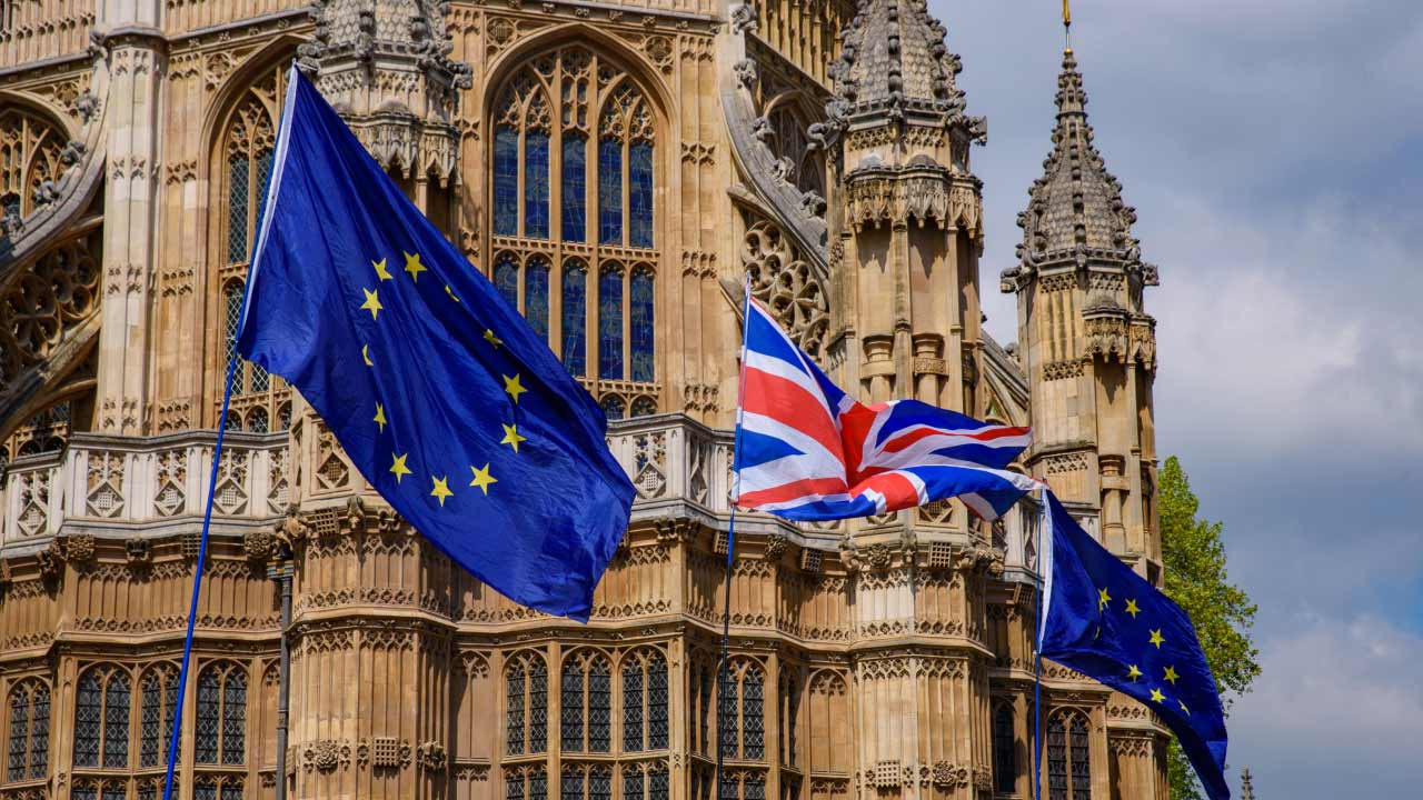 European Union flags and a Union Jack (United Kingdom flag) fly in front of a historic Gothic-style building, likely part of the UK Parliament in London, illustrating "How to Trade the Euro/British Pound?"