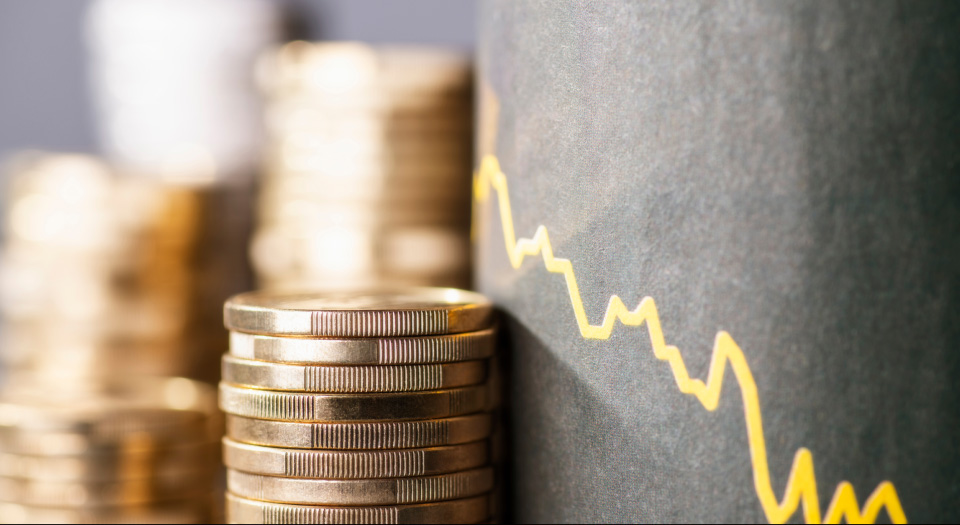 Stacks of gold coins next to a descending yellow line graph, symbolizing the impact of tariffs and immigration on inflation and economic decline