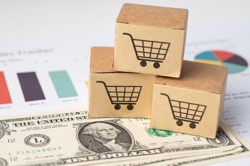 Stacked cardboard boxes with shopping cart icons placed on a US dollar bill, illustrating the concept of Forex Week Ahead Analysis: 20th January.