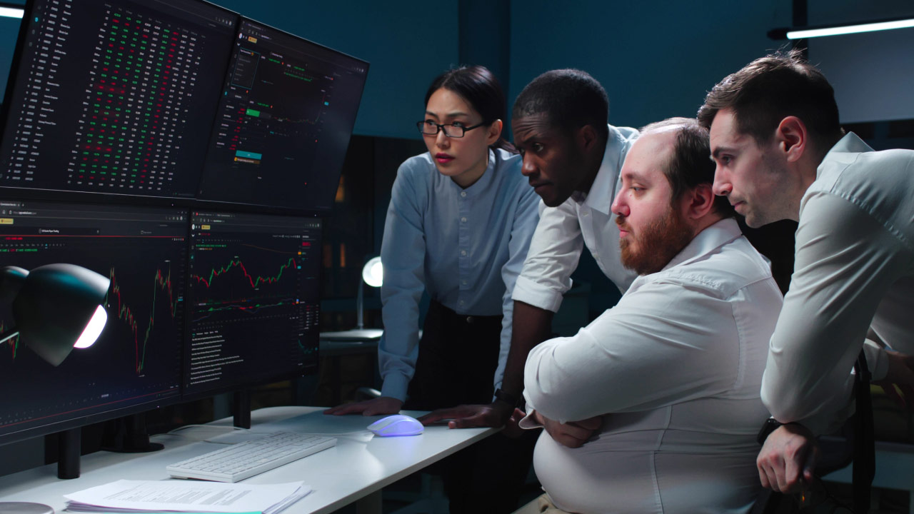 a group of four people, likely financial analysts or traders focused on multiple computer screens