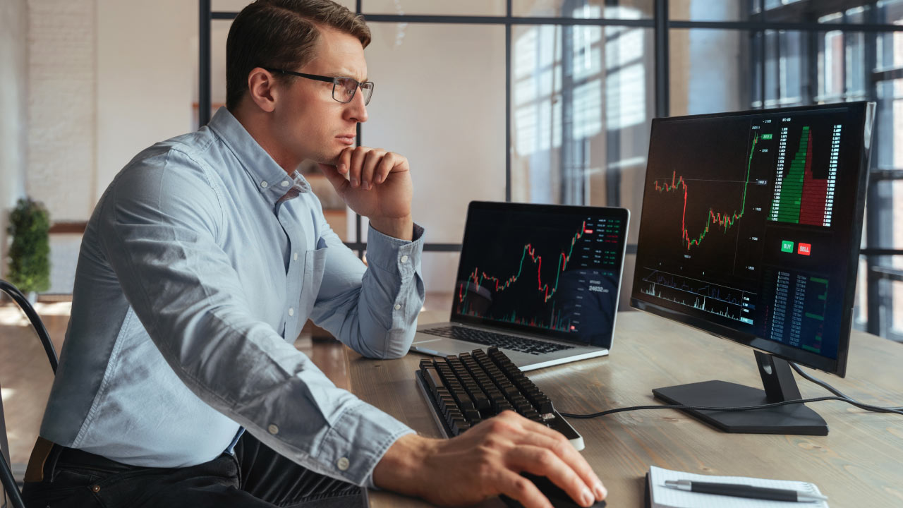 A focused man wearing glasses and a light blue shirt analyzing financial charts on two monitors at a modern office desk, illustrating helpful tips for forex trading newbies.