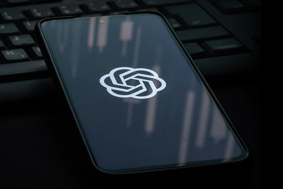 close-up of a smartphone displaying the ChatGPT logo, with a keyboard in the background and a faint candlestick chart visible on the screen.