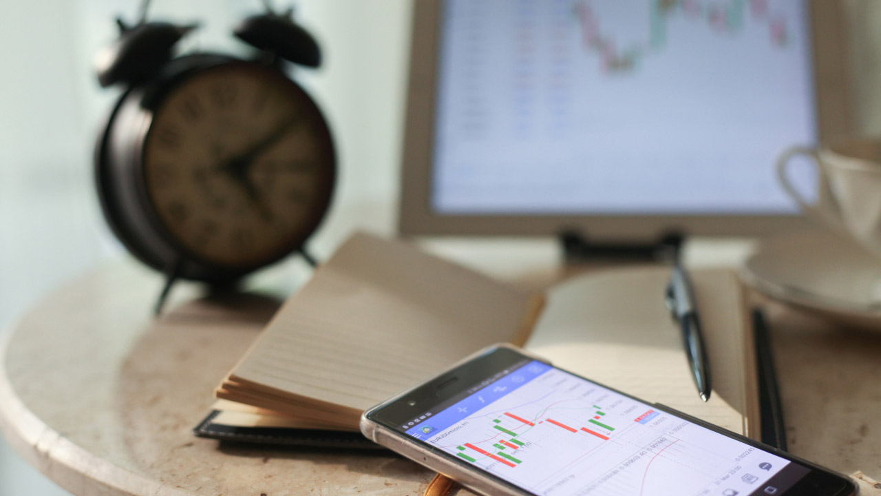 Smartphone showing candlestick charts beside a notebook, pen, and analog clock, representing the use of a trading journal.