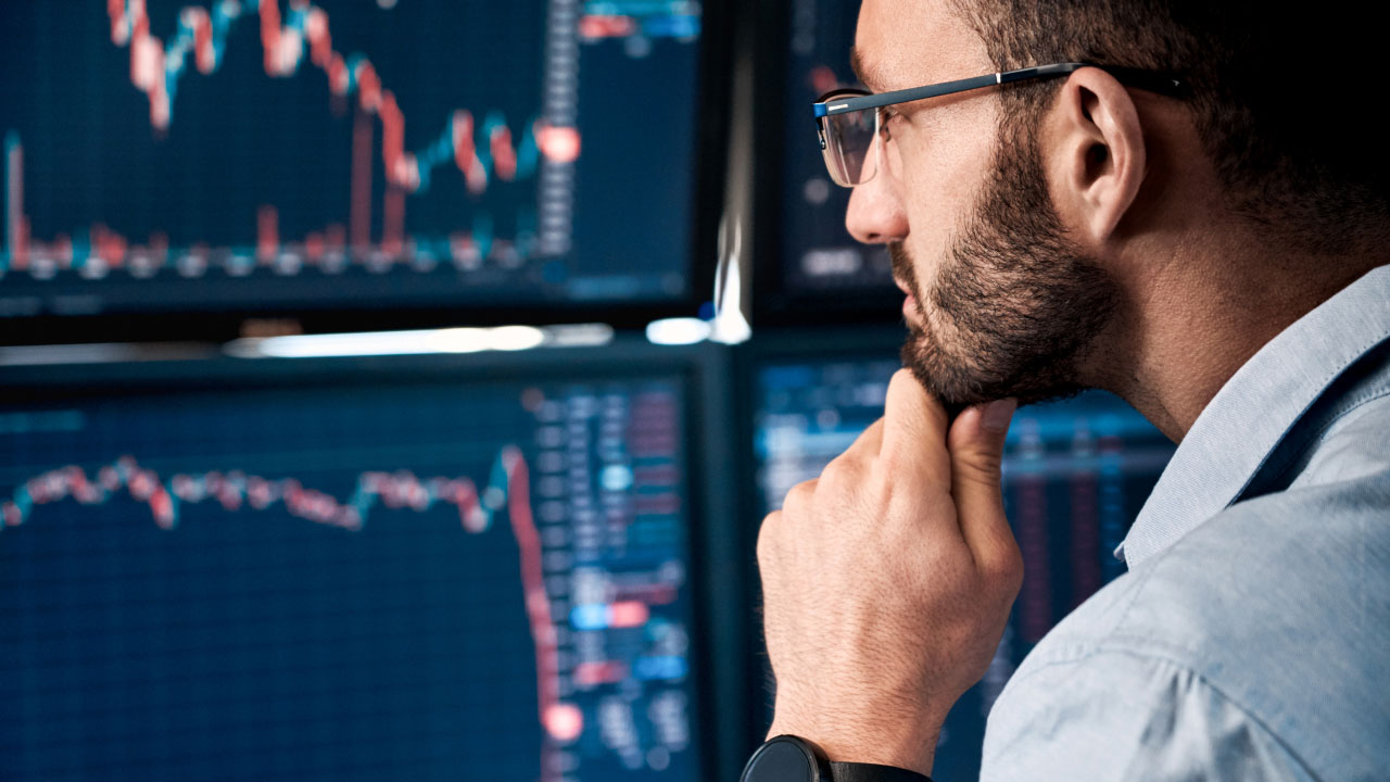 a man with glasses is intently studying multiple computer screens displaying financial charts 