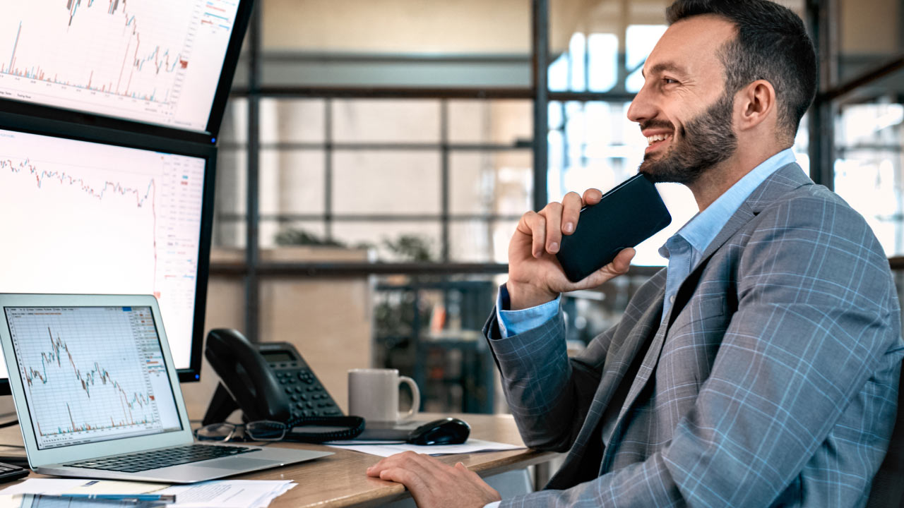 a smiling businessman in a suit holding a smartphone, sitting at his desk