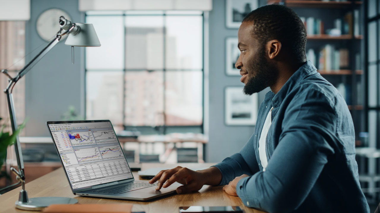 Man in a casual blue shirt sitting at a desk in a modern home office, illustrating how to transition from a demo account to a live account.