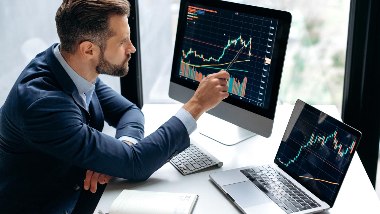 A professional trader in a suit analyzing financial charts on a desktop monitor and laptop, pointing at market trends with a pen. Top 8 Questions Introducing Brokers Need to Ask When Selecting a Broker.