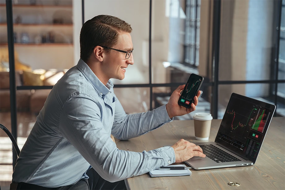 A man in glasses sits at a desk smiling while trading on both a laptop and smartphone, with financial charts displayed on the screens.