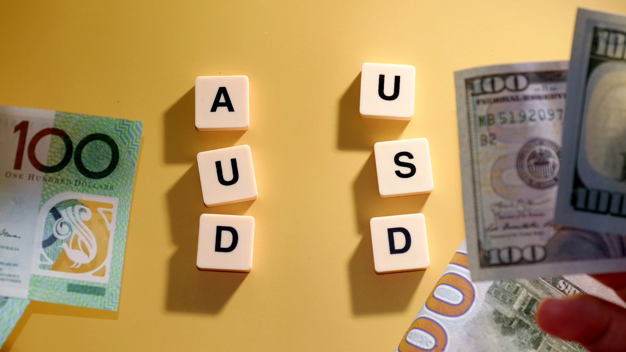 A visual representation of the AUD/USD currency pair. On a yellow background, the letters "AUD" and "USD" are arranged vertically using white letter tiles, illustrating the concept of "How to Trade AUD/USD."