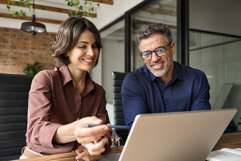 Two colleagues smiling while looking at a laptop screen during a discussion in a modern office setting.