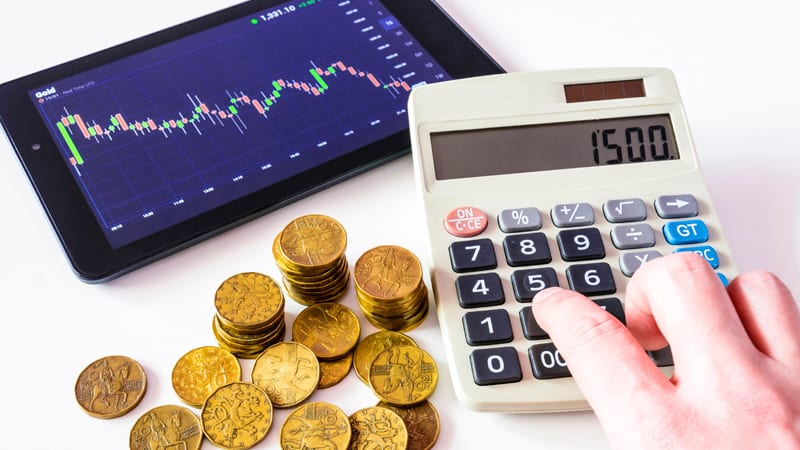 a calculator, stack of golid coins, and tablet showing a trading