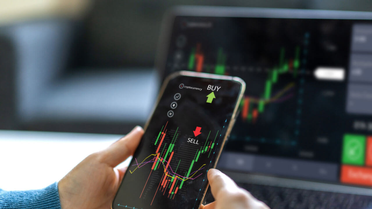 a person holding a smartphone displaying a trading app with "Buy" and "Sell" options