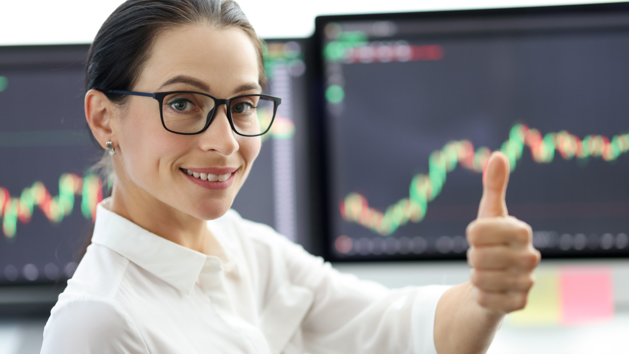 a confident female trader in glasses giving a thumbs-up in front of multiple monitors 