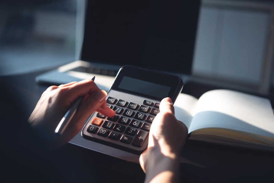Person using a calculator with a pen in hand, working at a desk with an open notebook and a laptop in the background.